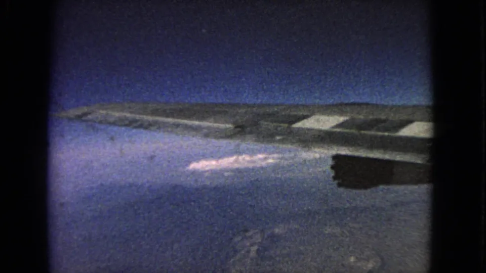1963 8mm passenger-window view with the aircraft wing and engine above blue haze, scattered clouds, and indistinct terrain or water.