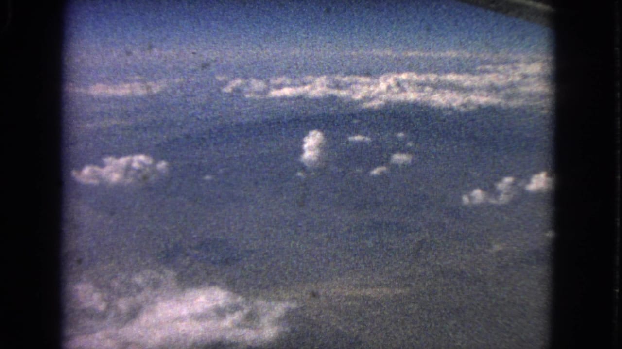 1963 8mm airplane-window view with blue sky, scattered white clouds, a distant haze line, and dark film gate edges.