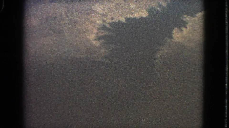 Final 1963 airplane-window frame showing clouds, heavy film grain, and a dark irregular shape that may be distant terrain, water, or shadow.