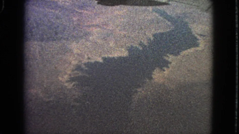 Supported 1963 home-movie aerial frame showing a dark wing-like shadow across grainy land and water near Barrington, Illinois.