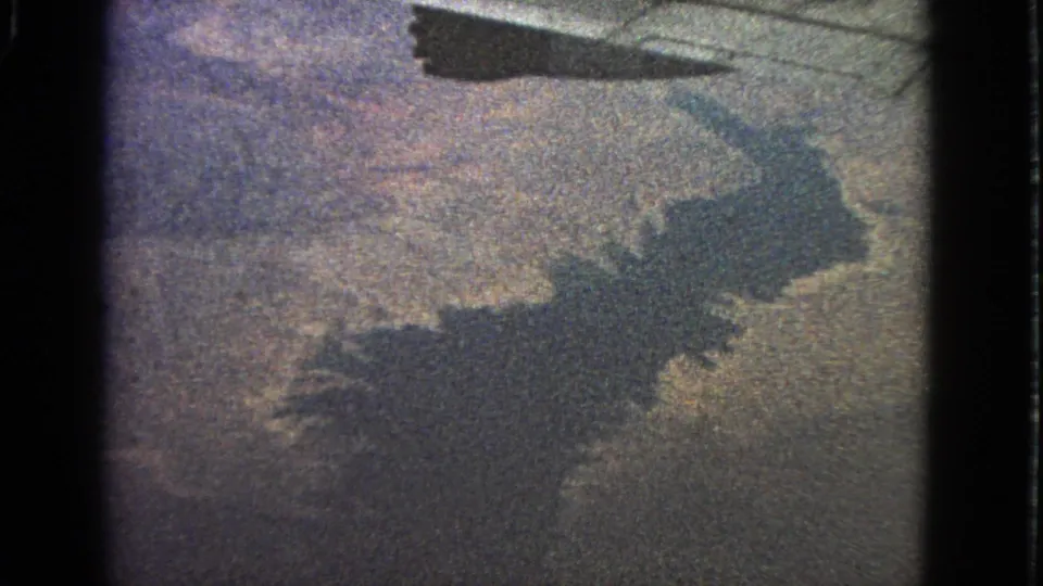 1963 8mm airplane-window view with a visible wing edge and dark shadow over muted land and water-like terrain.