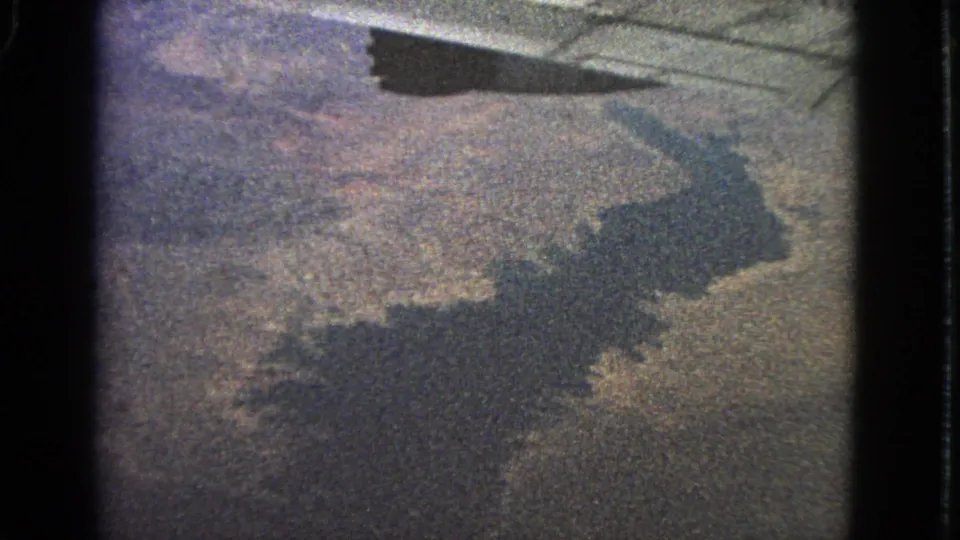 1963 8mm airplane-window view near Barrington, Illinois, showing a dark aircraft shadow over textured land and water.