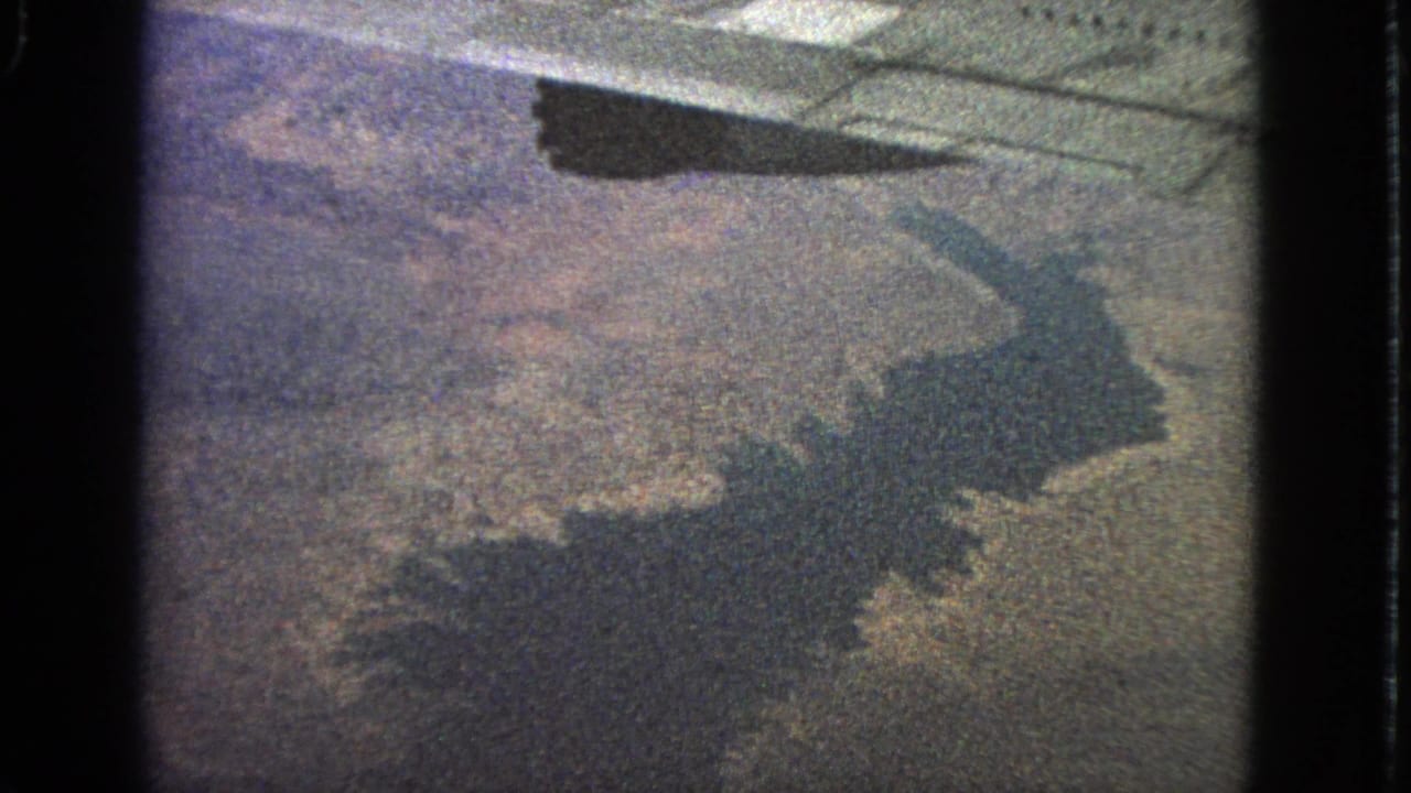 Plane shadow over mottled ground and water beneath an aircraft wing.
