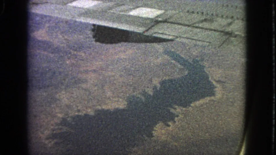 From a 1963 airplane window, the aircraft shadow falls across a grainy view of land and water, with the wing visible above.