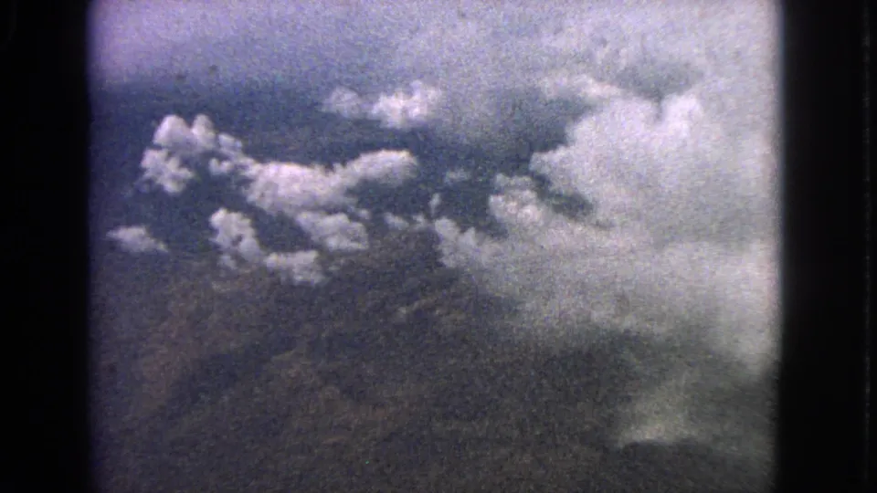 Cataloged 1963 8mm frame of clouds and hazy terrain below, filmed from inside an airplane.
