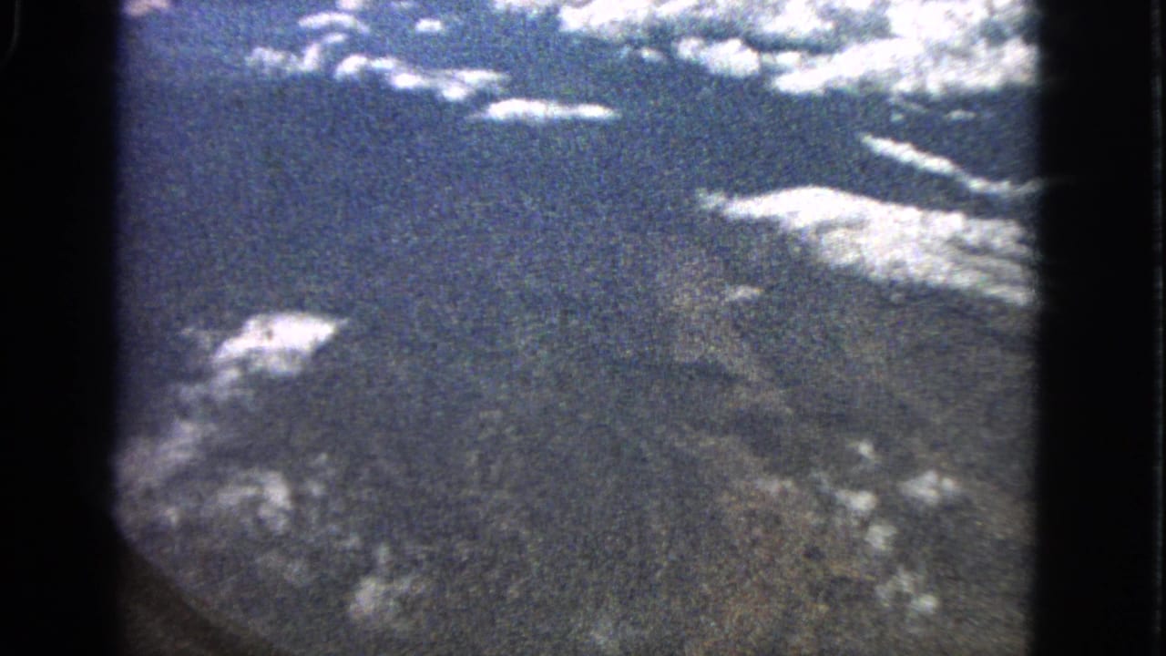 Aerial view through a dark airplane window edge with clouds over distant landforms.