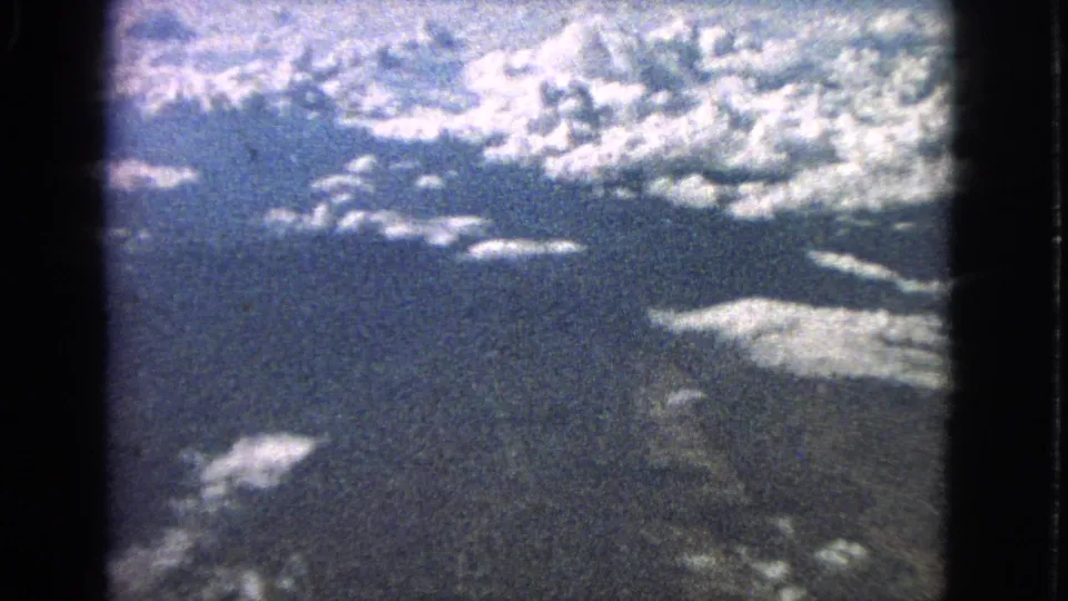 1963 8mm airplane-window view showing broad cloud bands over darker landforms with pronounced film grain.
