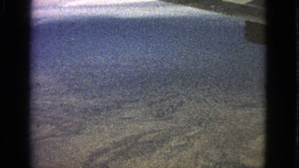 A passenger-view aerial shot shows distant ground texture and blue sky, with dark window or film-gate borders visible at the sides.