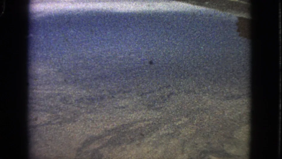 The 8mm frame holds on a hazy aerial landscape below a blue sky, viewed through a dark-edged aircraft-window composition.