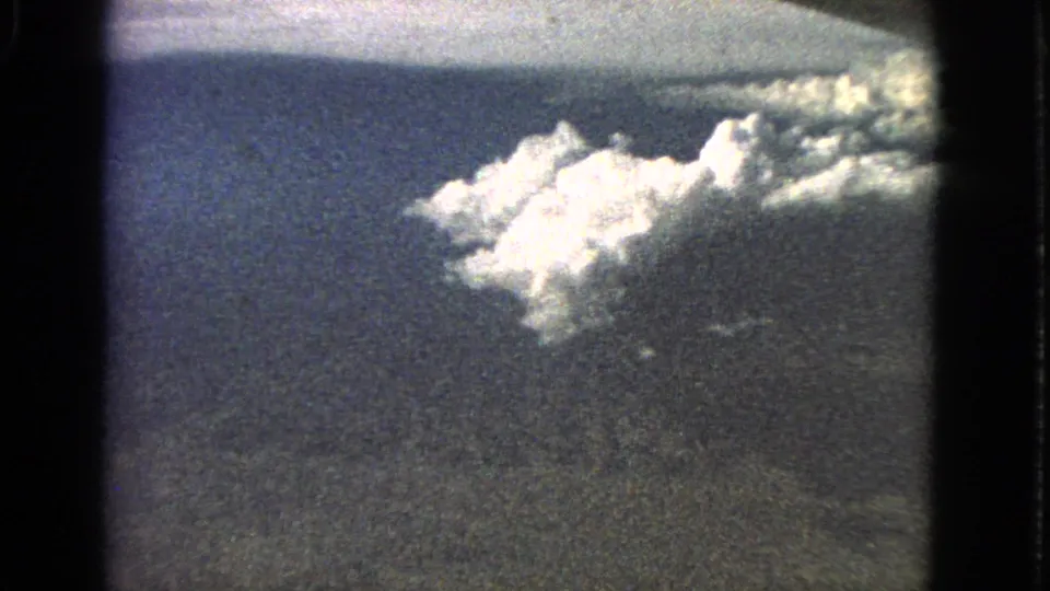 A bright cloud or plume-like formation appears above darker terrain in a grainy 1960s 8mm aerial frame from a plane window.