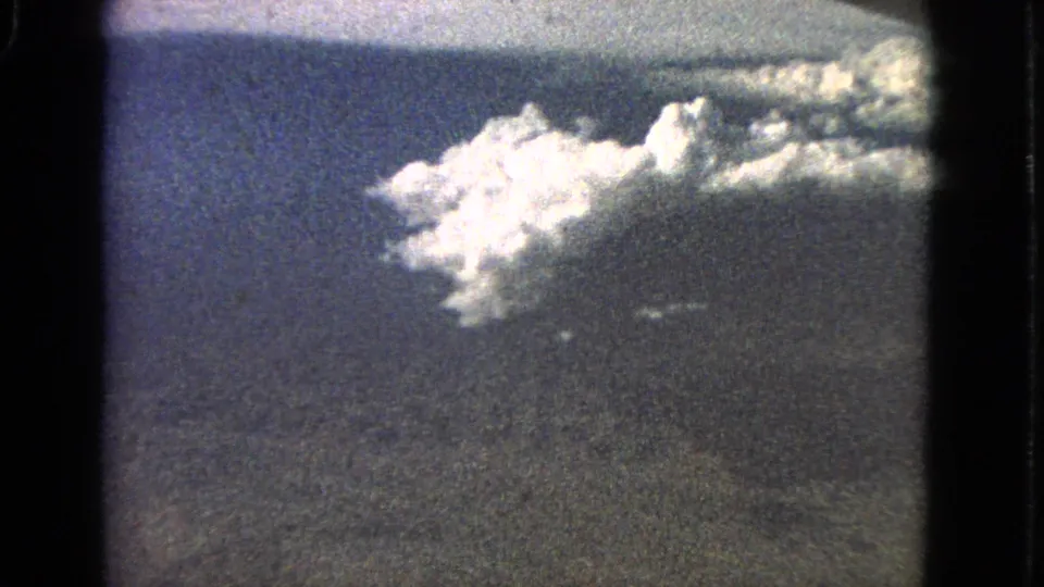 1963 8mm aerial frame linked to Barrington, Illinois, with bright clouds crossing a blue sky over dark landscape below.