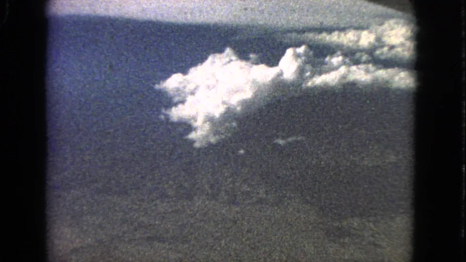 1963 8mm home-movie view from an airplane window, showing blue sky, a bright cloud formation, and a faint horizon.