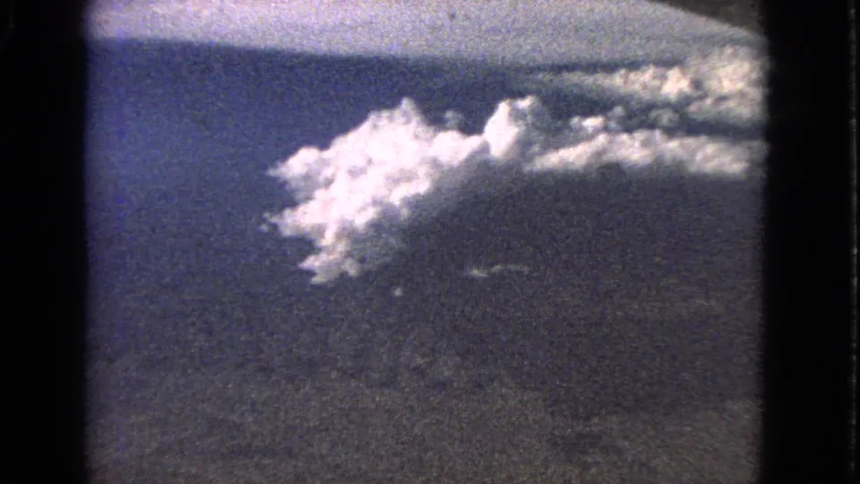1963 8mm airplane-window perspective with luminous clouds, blue sky, and visible film-frame borders.