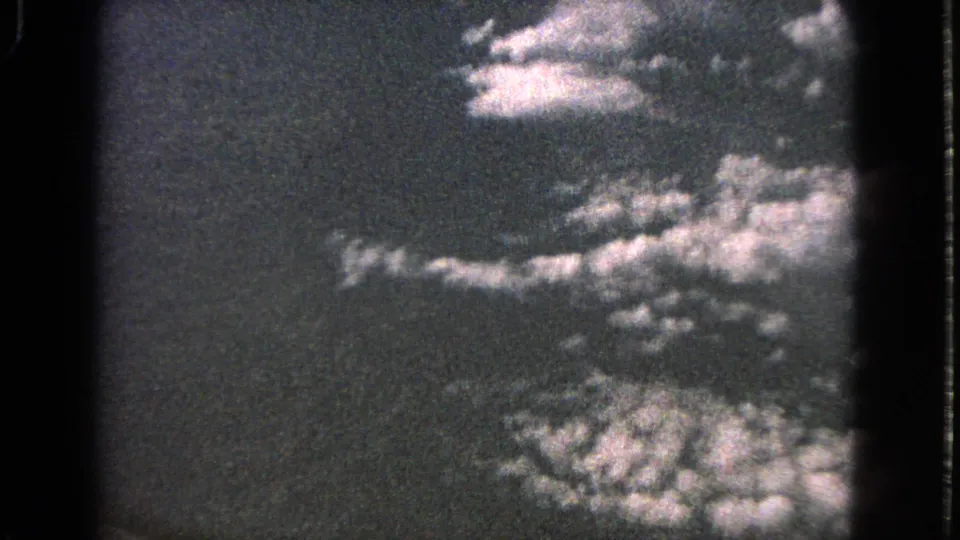 1963 8mm aerial frame with bright cloud clusters, dark sky, and indistinct lower texture from a passenger viewpoint.