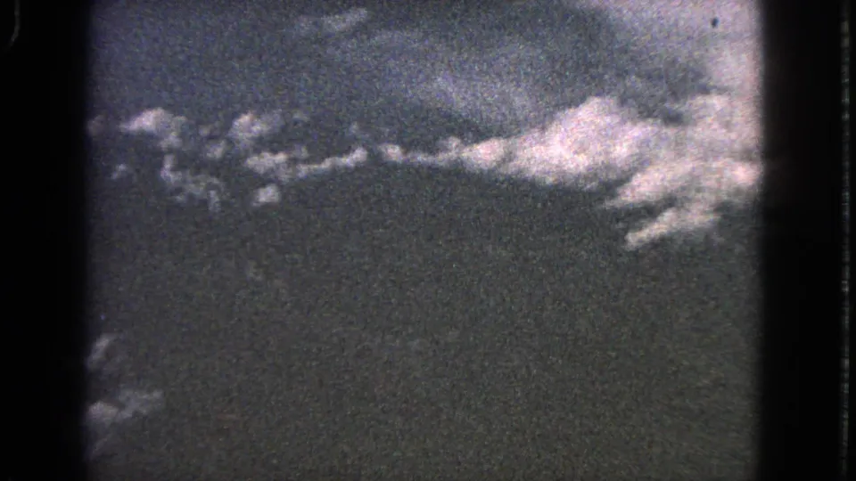 1963 8mm frame with a pale cloud mass and scattered cloud fragments seen from an airplane window.