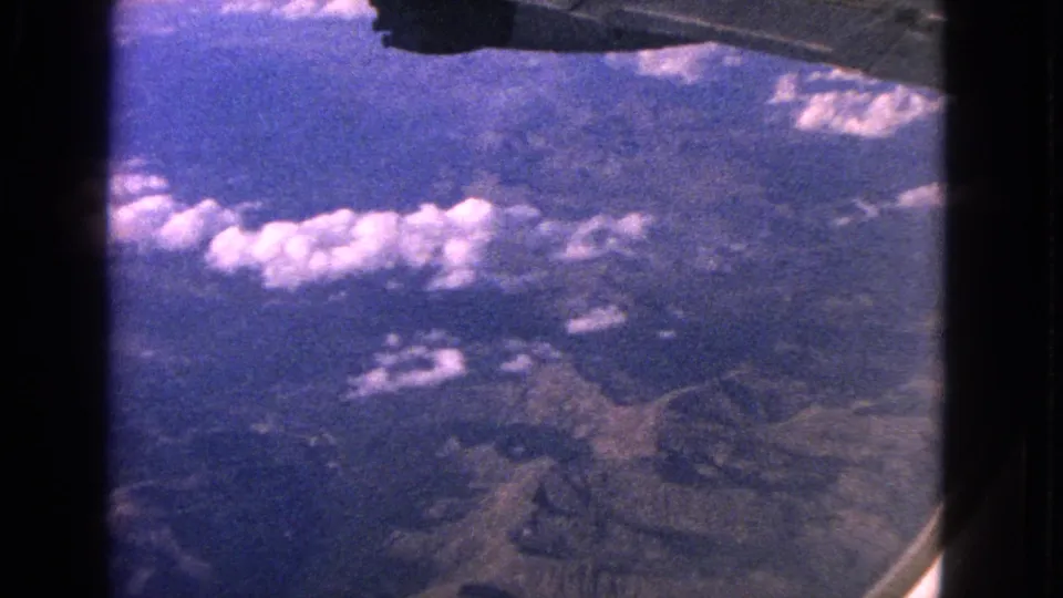 1963 8mm frame looking from a plane over rugged terrain, with clouds below and wing structure near the top.