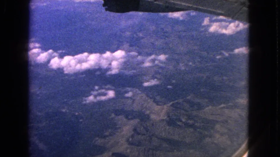 1963 8mm home-movie view from an airplane window, showing mountains, scattered clouds, and the aircraft wing edge.
