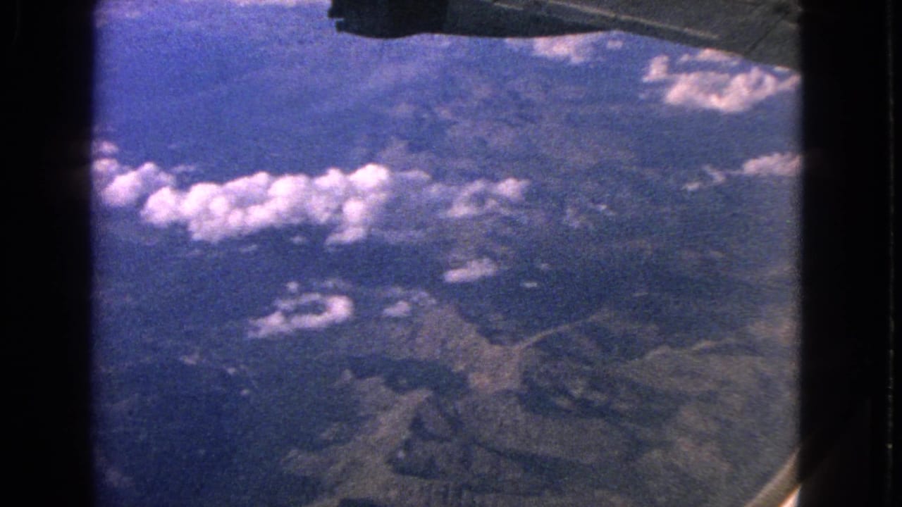 1963 8mm airplane-window view showing rugged mountain terrain, scattered clouds, blue haze, and part of the aircraft wing.