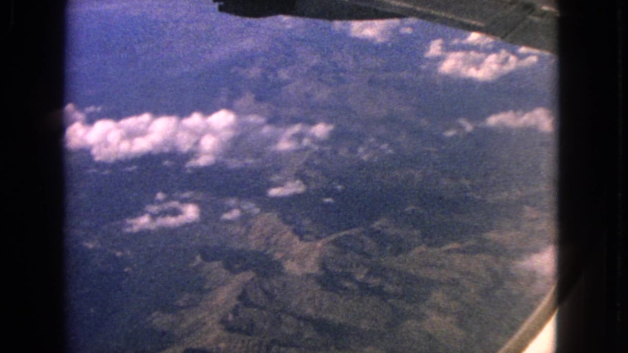 1963 8mm aerial view with the airplane wing across the top and hazy mountain terrain beneath scattered clouds.