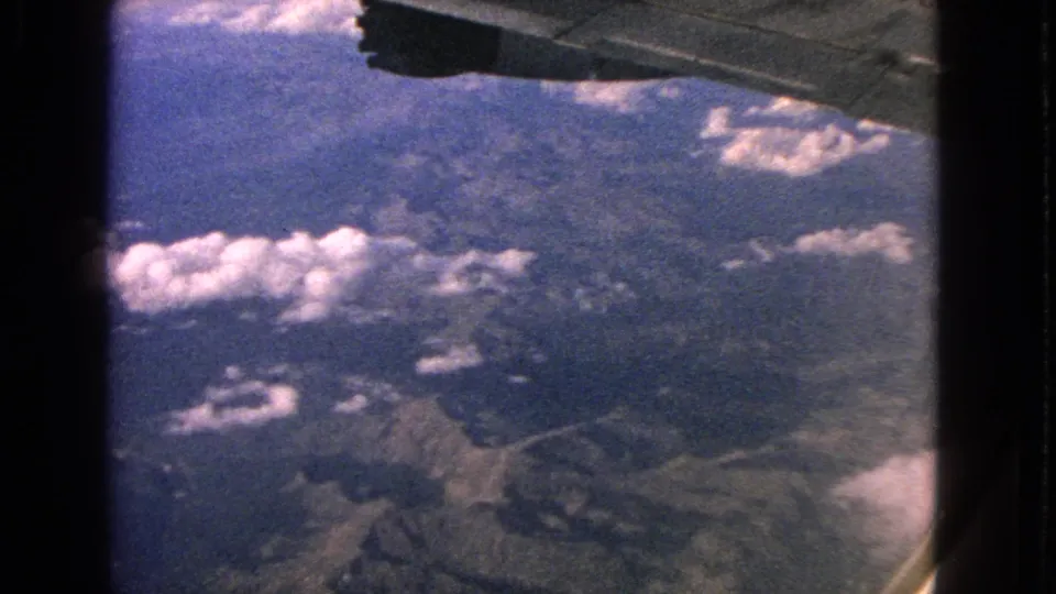 1963 8mm passenger-view footage showing mountainous terrain, bright clouds, and aircraft structure near the frame edge.
