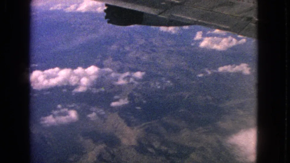 1963 8mm aerial frame showing a hazy mountain landscape, scattered clouds, aircraft wing detail, and dark film-edge shadows.