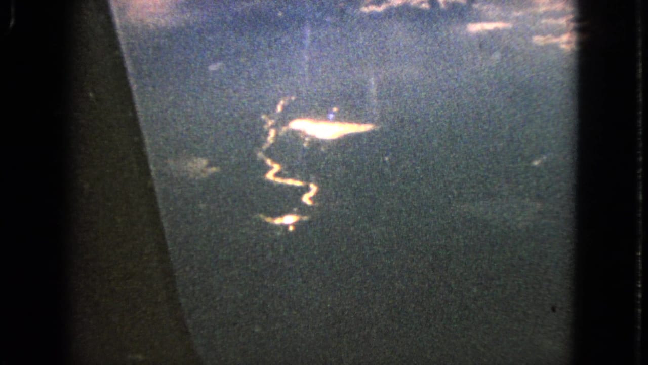 1963 8mm plane-window view over water, with sun glare on the surface and dark window framing at the side.