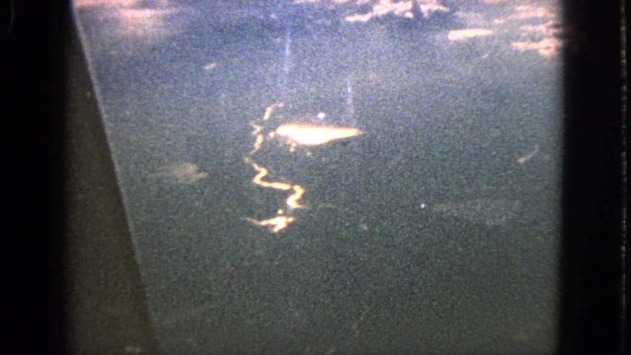 1963 8mm aerial view from a passenger window, showing a bright sun streak across dark water beneath clouds.