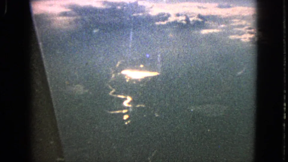 1963 8mm plane-window view with bright reflected sunlight on water and cloudy sky texture near the top of frame.