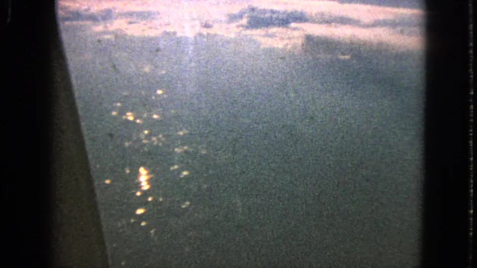 A 1963 8mm airplane-window frame shows hazy terrain or water below with scattered sun reflections and cloudy sky at the top.
