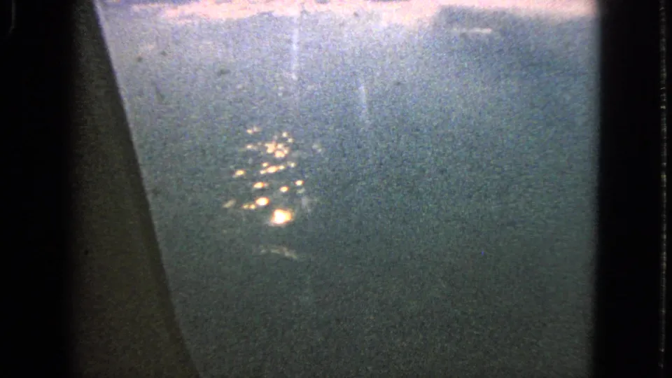 A grainy 1963 8mm frame shows bright clusters of night lights seen from an airplane window over an indistinct dark surface.