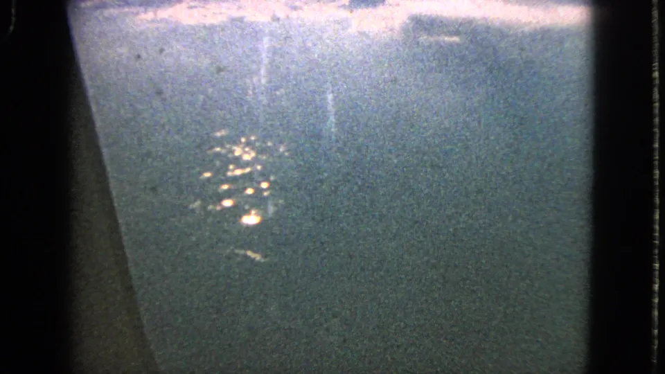 1963 home-movie frame from an airplane window, with clustered lights below and a dark window edge cutting through the grainy night view.