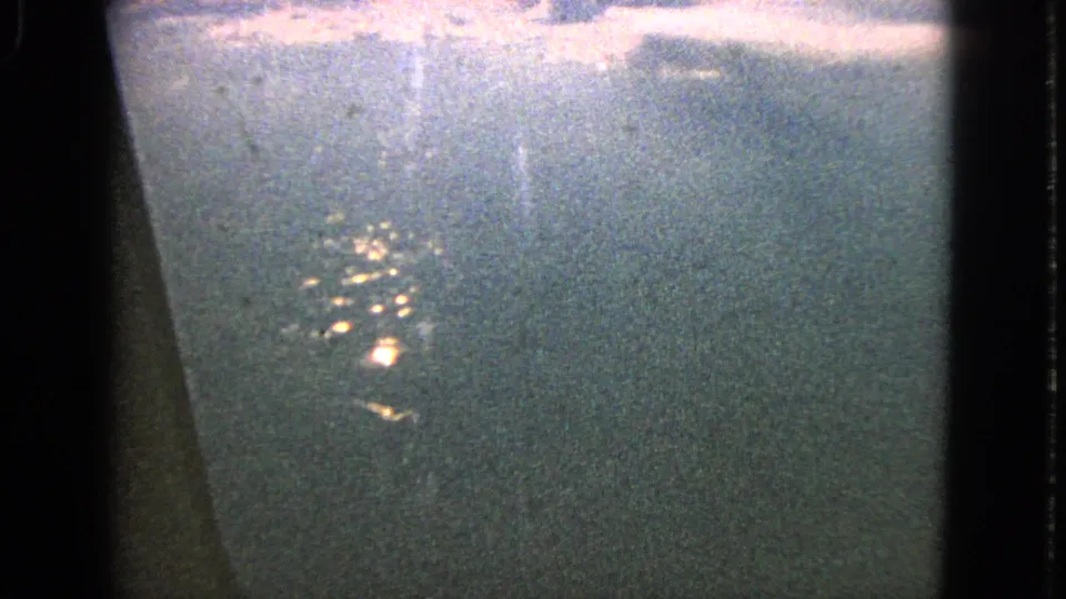 A 1963 8mm airplane-window shot near Barrington shows a small cluster of night lights below, softened by grain, haze, and window glare.