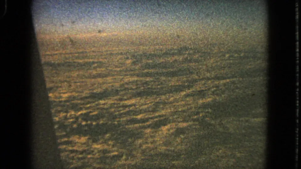 1963 8mm airplane-window view with a dark window edge and textured cloud cover below a hazy sky.