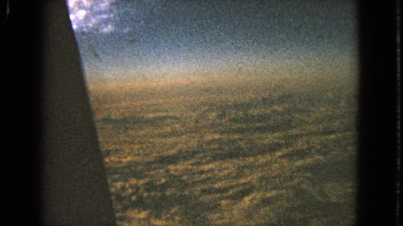1963 8mm frame looking past a dark airplane-window edge toward hazy sky and distant ground during approach or descent.