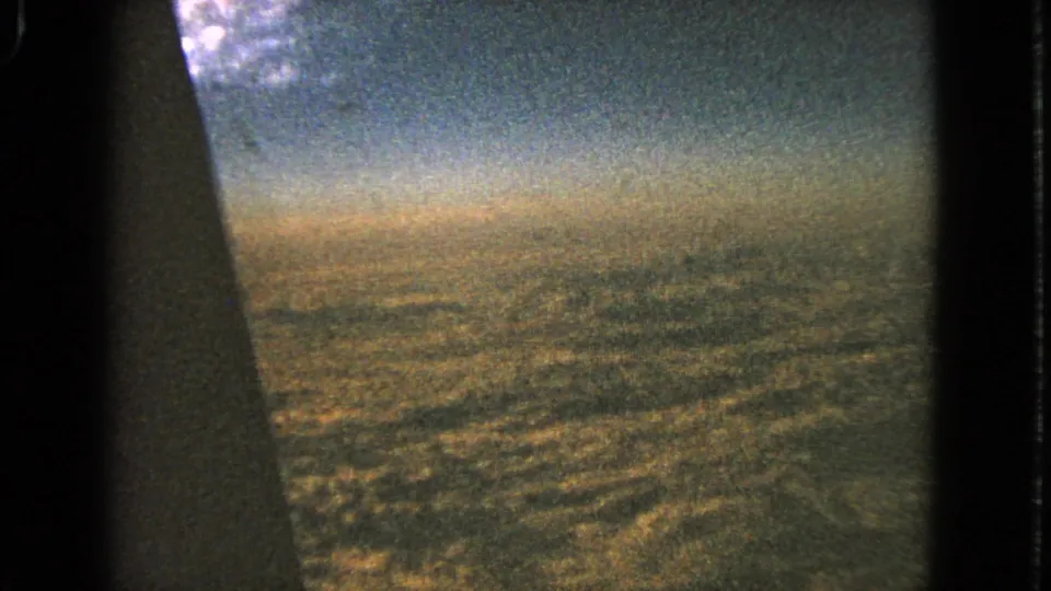 1963 8mm frame looking past a dark airplane-window edge toward hazy sky and distant ground during approach or descent.