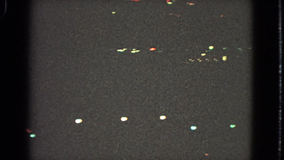 Grainy 1963 home-movie frame with scattered Christmas lights and soft reflections during a nighttime drive.
