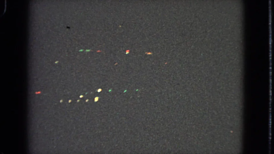 Nighttime airplane-window footage on 8mm film, showing faint colored lights below and heavy low-light grain.