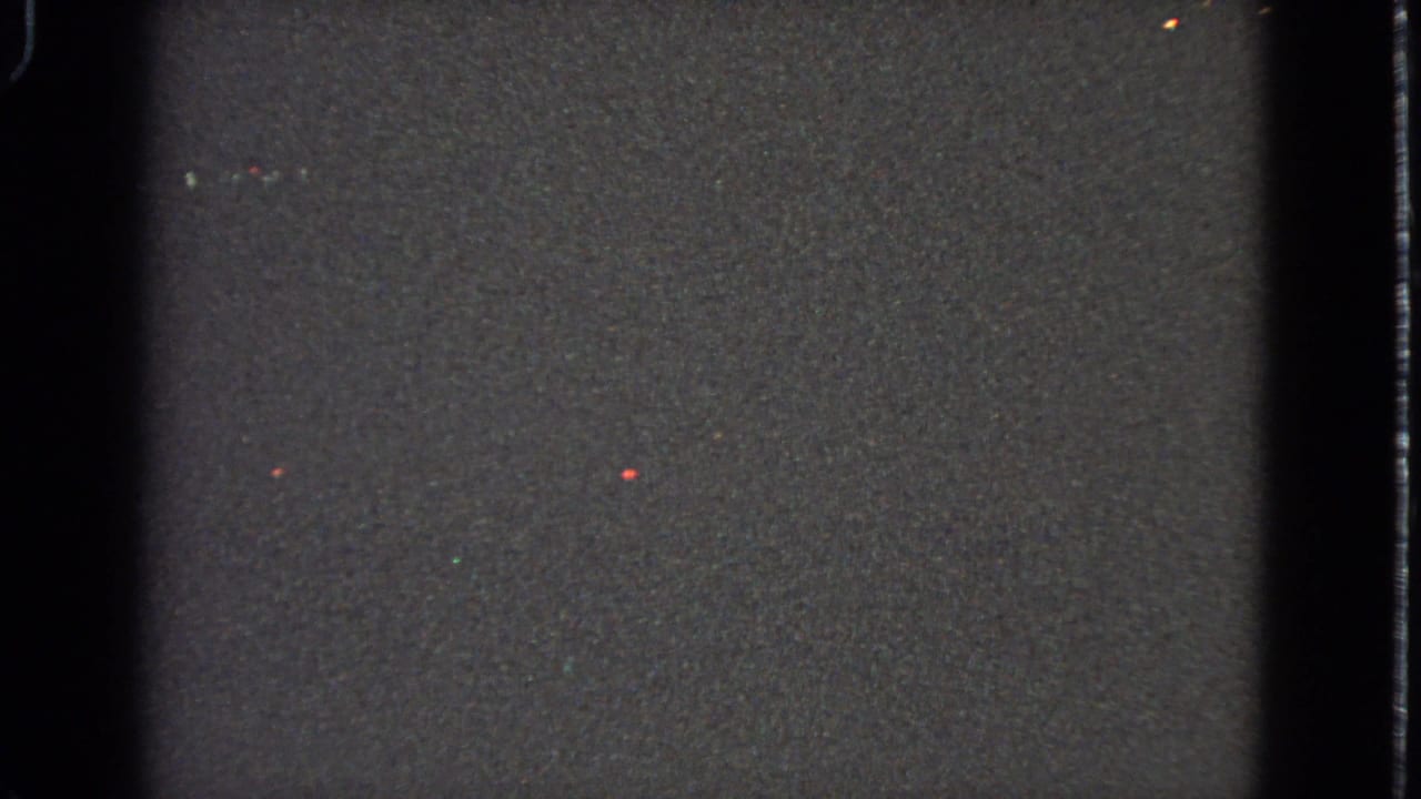 Dark grainy film frame with vertical borders and faint colored specks.