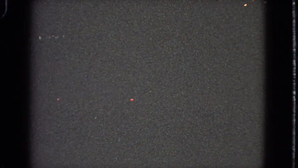 Another dark 8mm frame with visible grain, black frame edges, and faint colored specks from the scanned film.