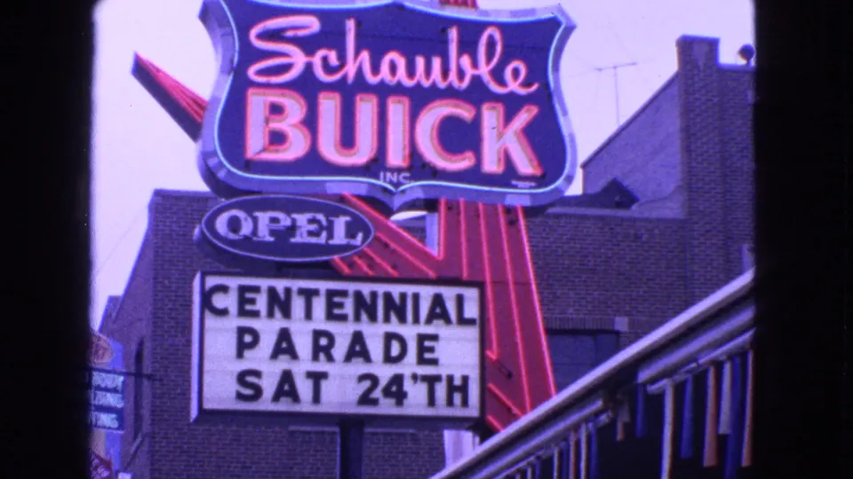 1963 Barrington view of a neon Schaubie Buick sign, Opel logo, and marquee reading Centennial Parade Sat 24th on a brick building.