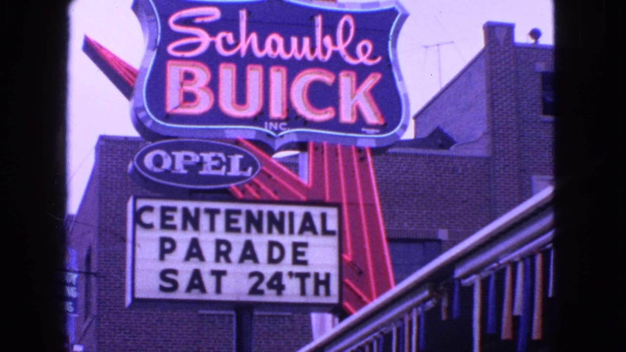 1963 Barrington, Illinois frame showing Schauble Buick neon, an Opel sign, and a Centennial Parade marquee on a brick commercial facade.