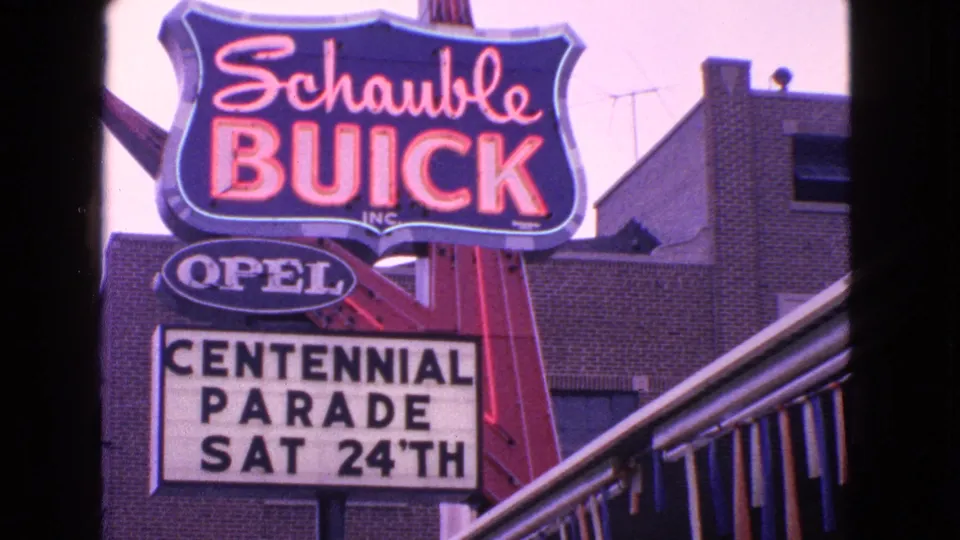 1963 Barrington, Illinois frame showing Schaubie Buick and Opel signage with a Centennial Parade marquee on a brick storefront.