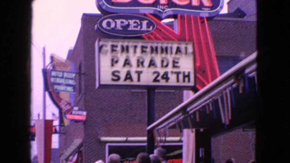 1963 Barrington, Illinois street view with the Centennial Parade marquee, Opel sign, storefront awning, and adults gathering below.