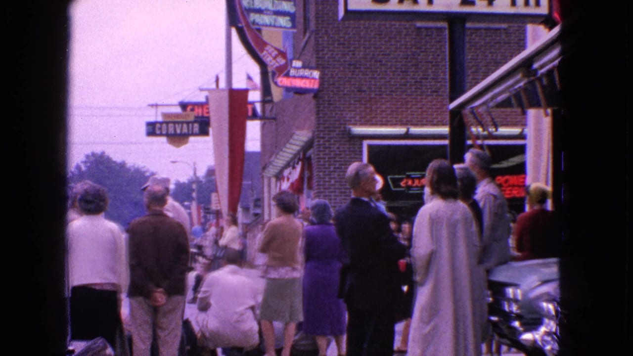 Adults in 1963 Barrington, Illinois stand along a commercial sidewalk near storefront and auto-related signs before a parade.