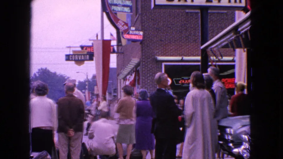 Adults in 1963 Barrington, Illinois stand along a commercial sidewalk near storefront and auto-related signs before a parade.
