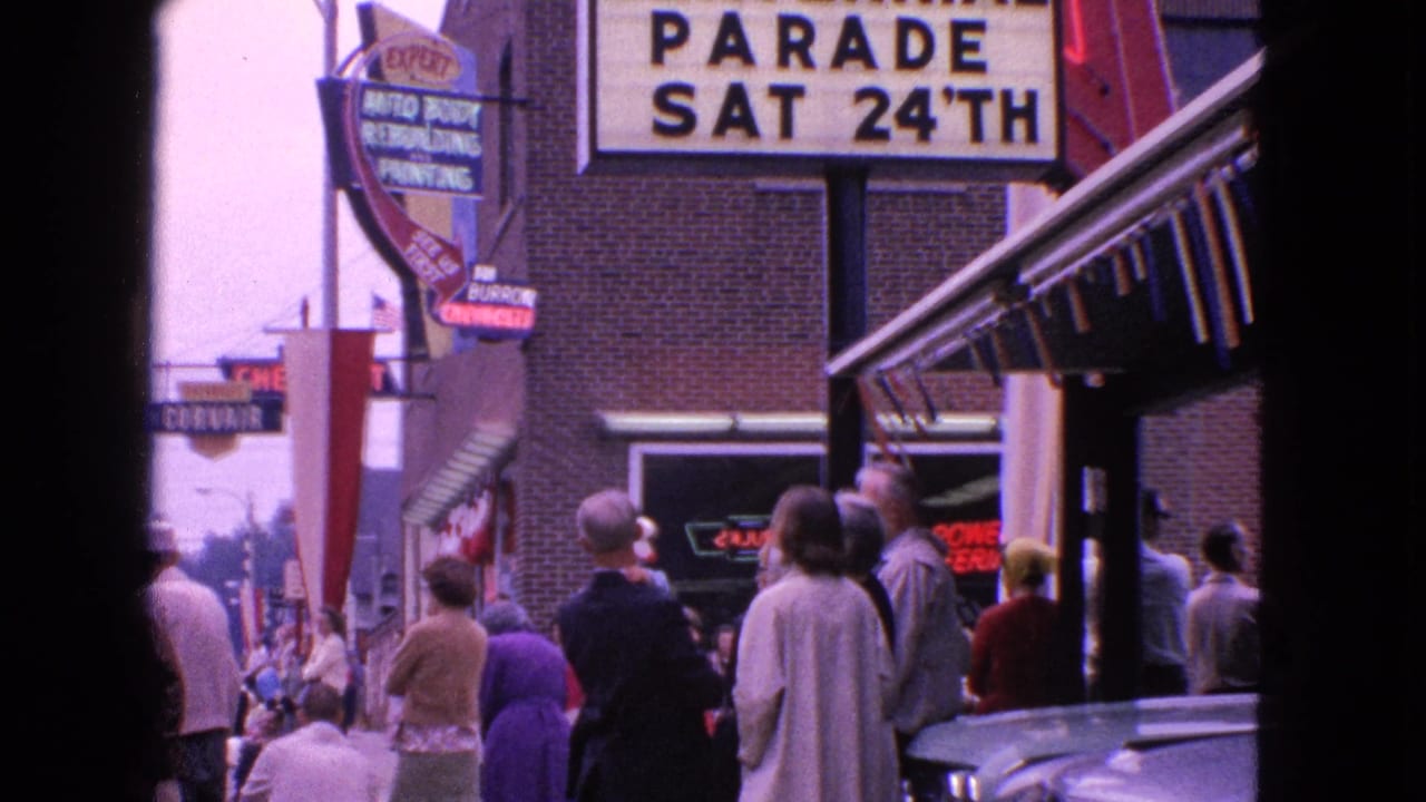 Barrington sidewalk spectators stand beneath a large parade announcement sign and mid-century storefront awnings.