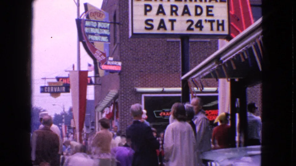 1963 Barrington street view with adults waiting on the sidewalk under a parade sign and commercial auto signage.