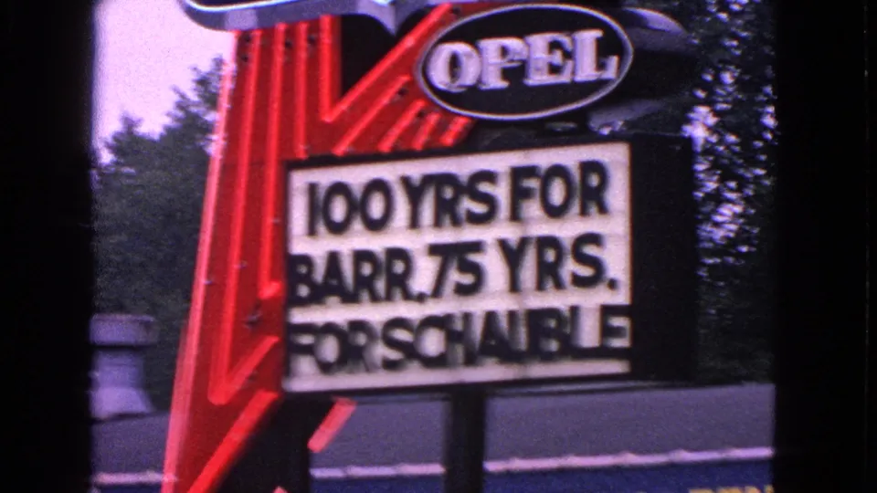 Final 8mm frame keeps the Opel sign and anniversary marquee in view, with partial yellow lettering visible below.