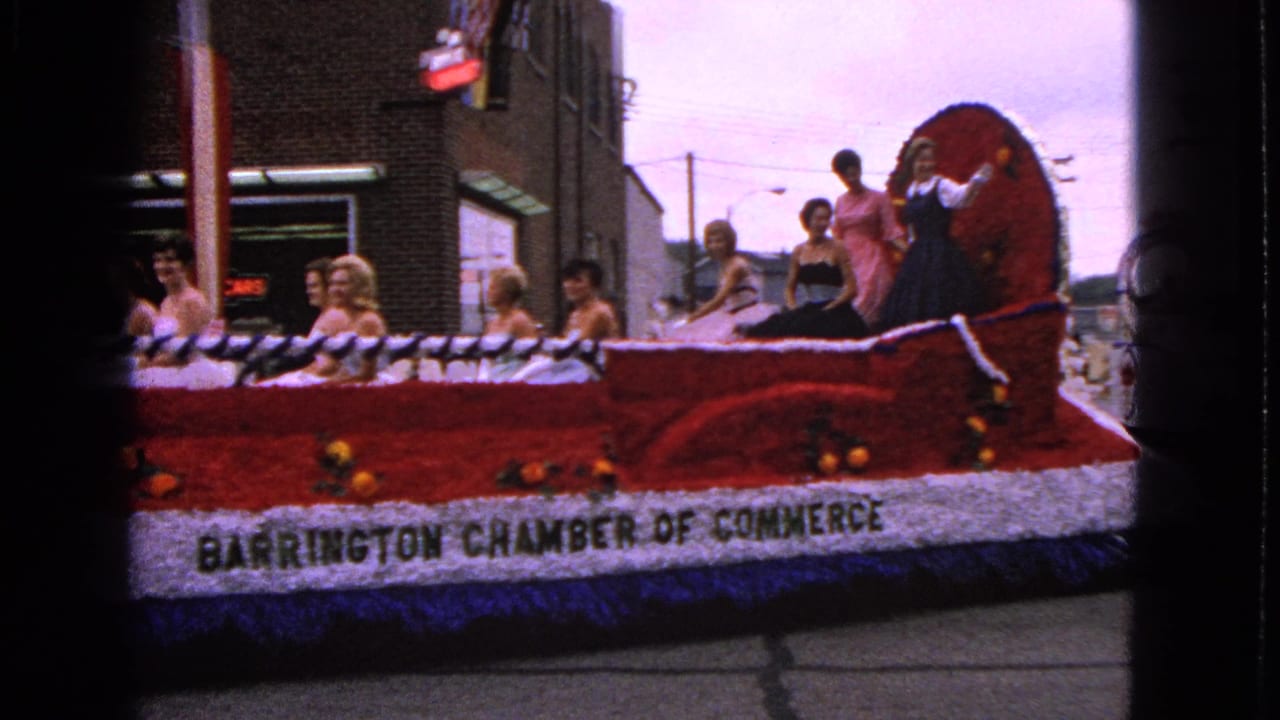 Red floral parade float carrying women in formal dresses past a brick storefront.