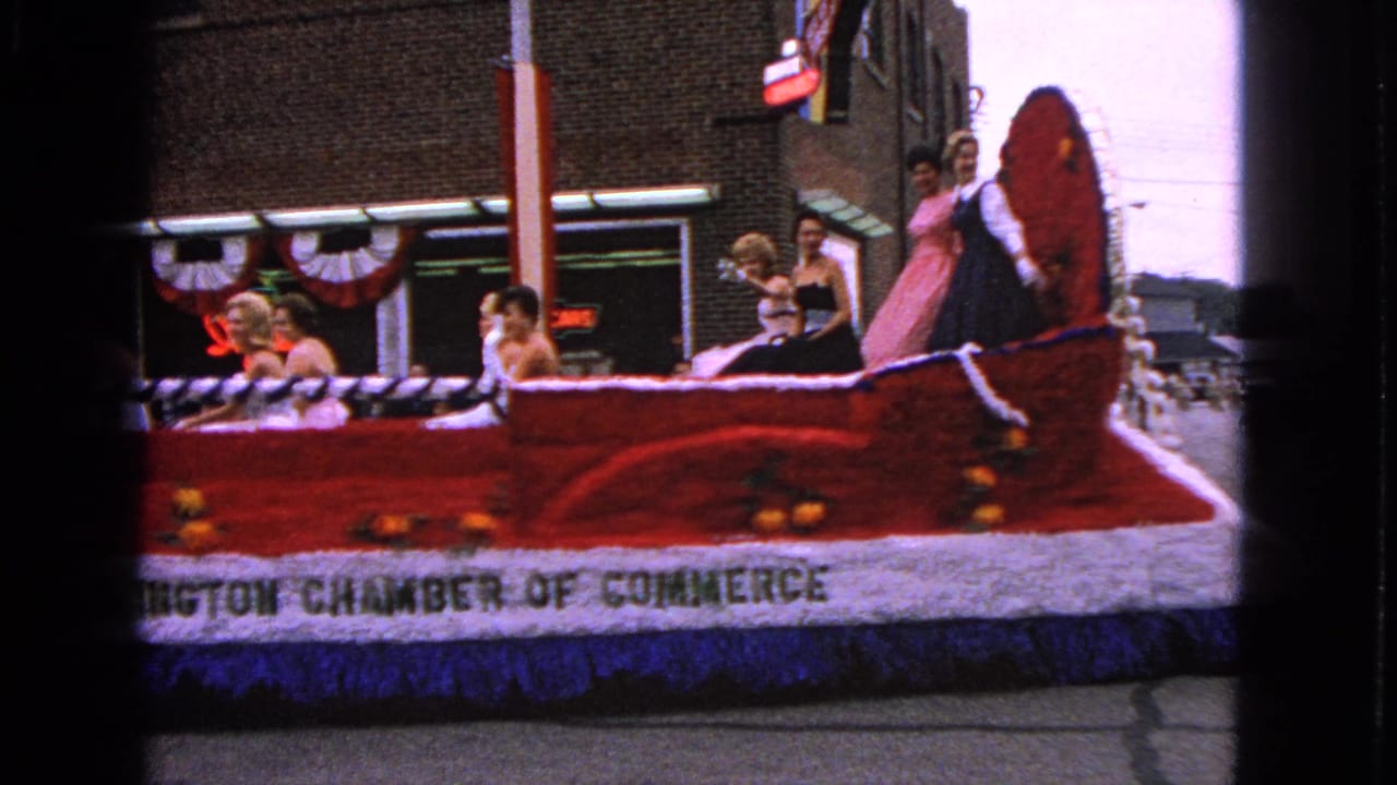 Side view of a red floral float with riders passing a decorated storefront.